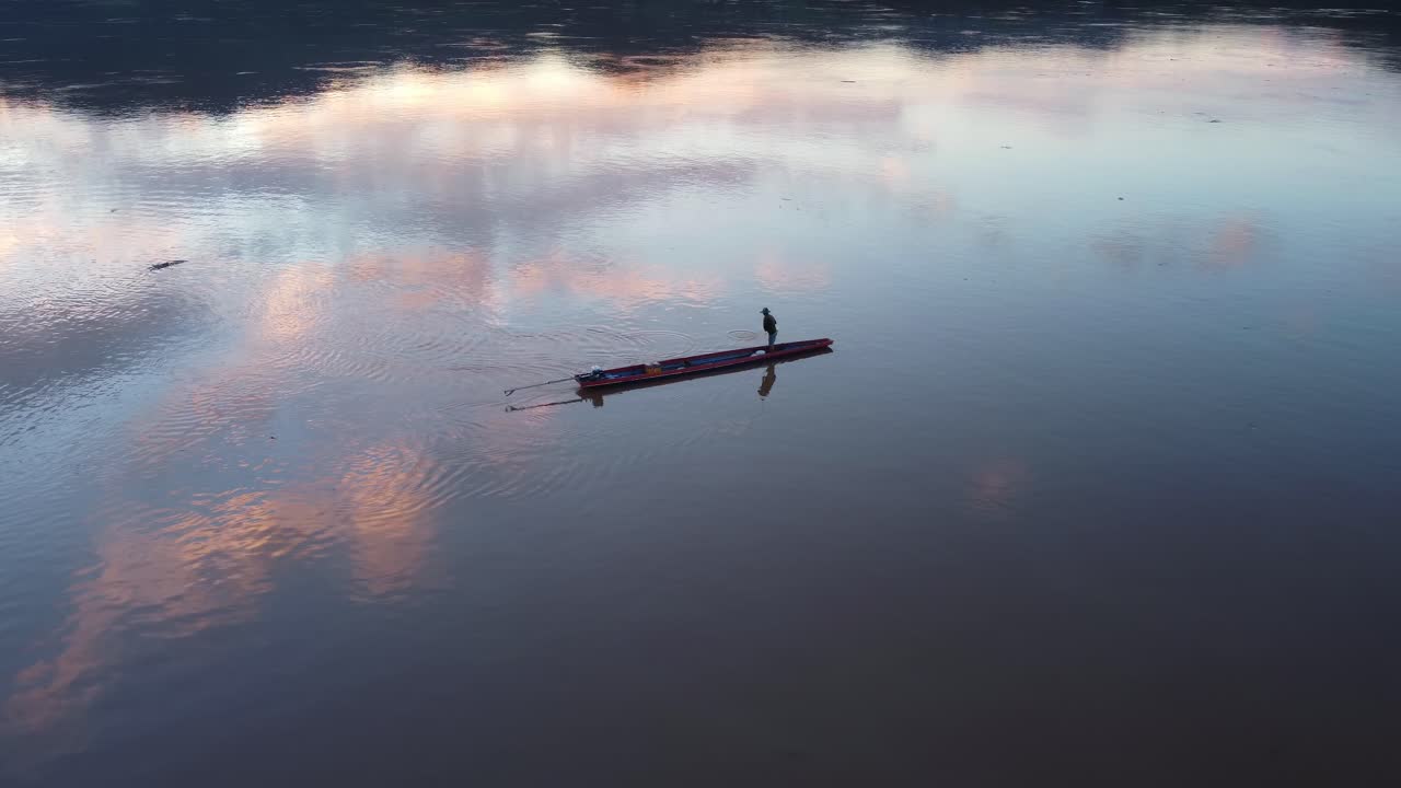 외로운 시이 은 롱테일 보트 위에 서서, 멋진 일몰 구름이 반사되는 하늘 아래 조용한 강에 그물을 던지고 있습니다.