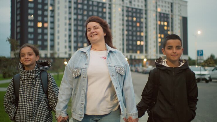 Smiling woman walks hand in hand with two cheerful children in urban residential area during evening, surrounded by tall apartment buildings, expressing joy, connection, and warmth in daily city life