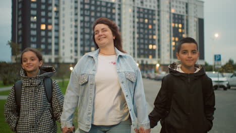 smiling woman walks hand in hand with two cheerful children in urban residential area during evening, surrounded by tall apartment buildings, expressing joy, connection, and warmth in daily city life