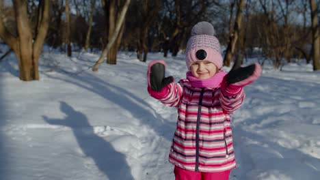 Child-girl-looking-at-camera,-smiling,-dancing,-waving-hands-on-snowy-road-in-winter-park-outdoors