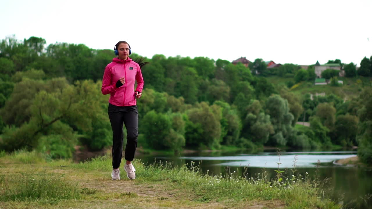 A girl in a pink jacket and black pants runs near the river in headphones preparing for the marathon