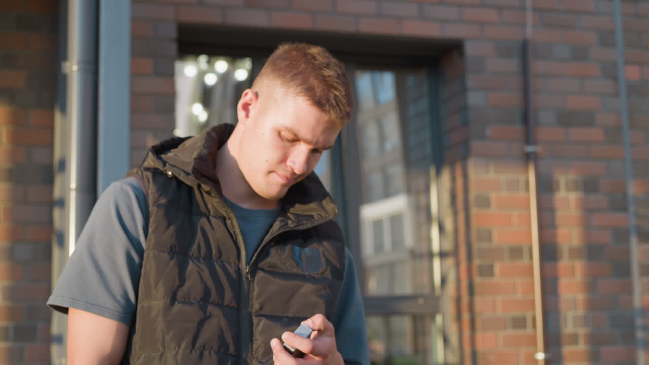 young man standing outside brick building under sunlight holds cigarette pack, makes decisive gesture to toss it into nearby waste bin and pauses reflectively after action, contemplating change