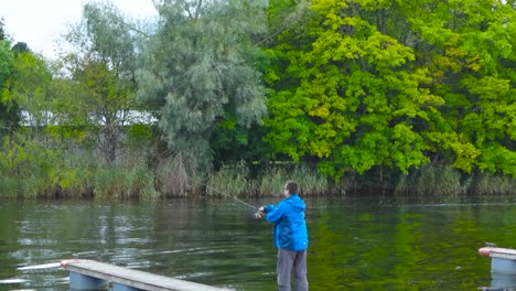 A man fishing with a spinning rod on a dock near a rural and small river that is surrounded by greenery and plants during summer. Water has ripples and reflects light. The man throws bait slow motion.