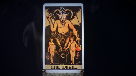 a tarot devil card sits on a brass stand as smoke rises around it