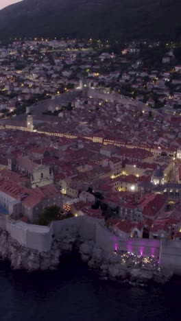 city walls protecting the old town of dubrovnik with its buildings illuminating at dusk, croatia. aerial, vertical video