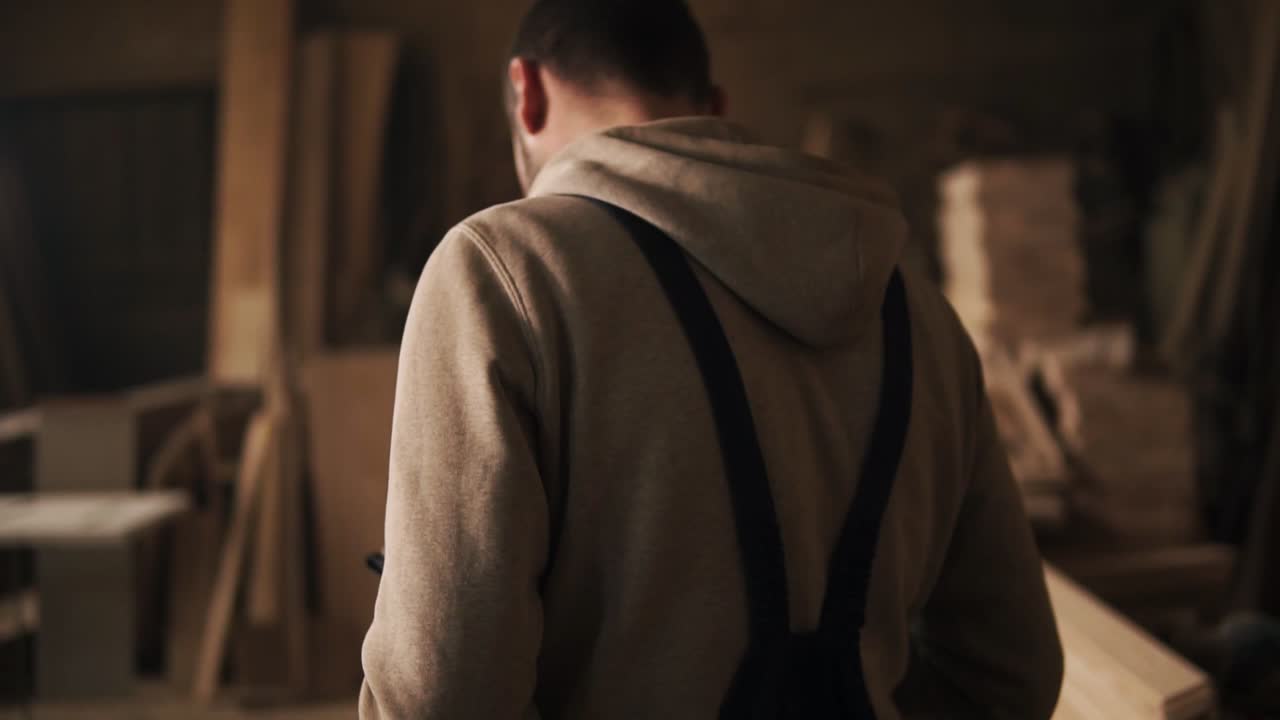 스웨트셔츠와 파란색 오버럴을 입은 젊은 남자가 목수 작업실을 가로질러 가는 뒷면의 모습. 가구. 느린 움직임