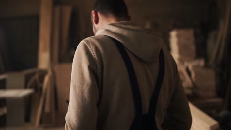 backside view of a young man in a sweatshirt and blue overalls going through a carpentry workshop. furniture. slow motion