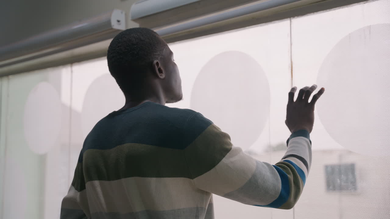 Man overlooking street through patterned blind. Back view in striped sweater, soft daylight and muted city skyline beyond glass, hand raised to veil, distant gaze suggesting waiting or missed date
