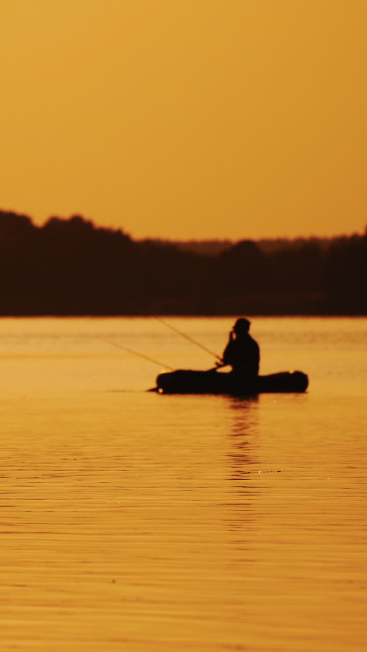 Golden sunset and the silhouette of a man throwing a fishing rod from the boat. Fisherman is catching fish from a rowboat in the evening on the background of natural landscape. Vertical video