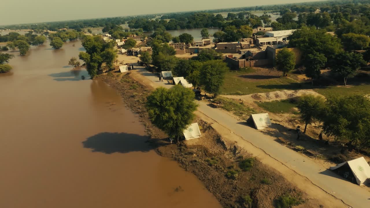 Low drone flight along dirt road separating flooded fields with improvised tents in dry land with houses under intense sun