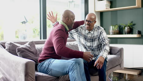 happy african american senior father and son sitting on couch,embracing and laughing, slow motion