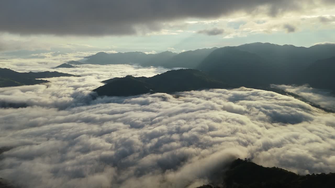 Wide aerial view of mountain ridges rising above a sea of clouds, with soft sunbeams and dramatic overcast sky creating a moody morning atmosphere in a remote natural landscape