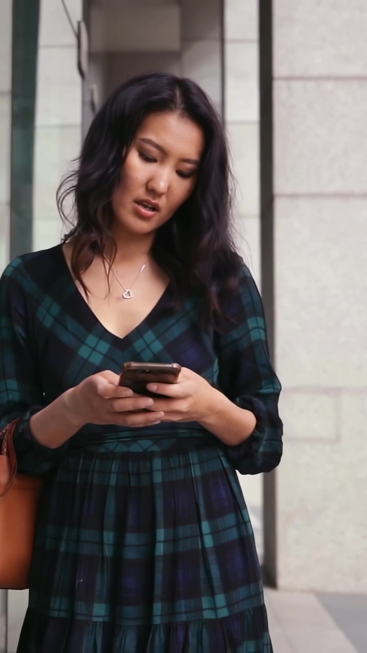 Video verticale: Donna che legge, con un vestito a quadri, digita sullo smartphone all'ingresso, tenendo una borsa di pelle.