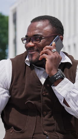 positive black skinned businessman having a phone talk in the city. african american man in glasses sitting on a bench near office center and speaking the phone. vertical video