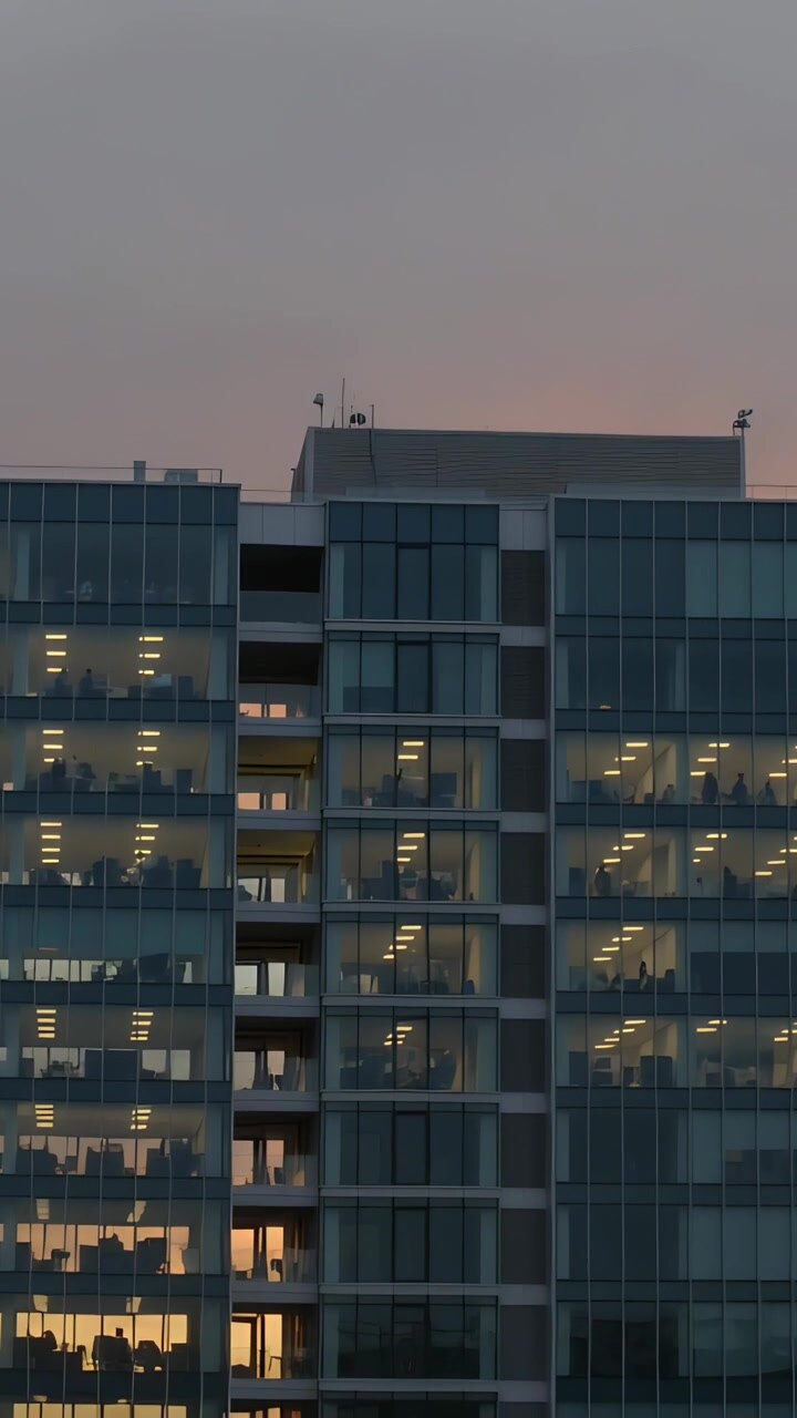 Vídeo vertical: Luz crepuscular mutável aquecendo a fachada de vidro de um escritório no centro da cidade, mostrando mesas e monitores.
