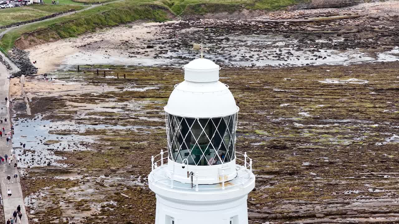 Drone camera smoothly ascends above St. Mary’s Lighthouse, revealing rocky tidal flats, a coastal causeway, and visitors walking below in soft daylight
