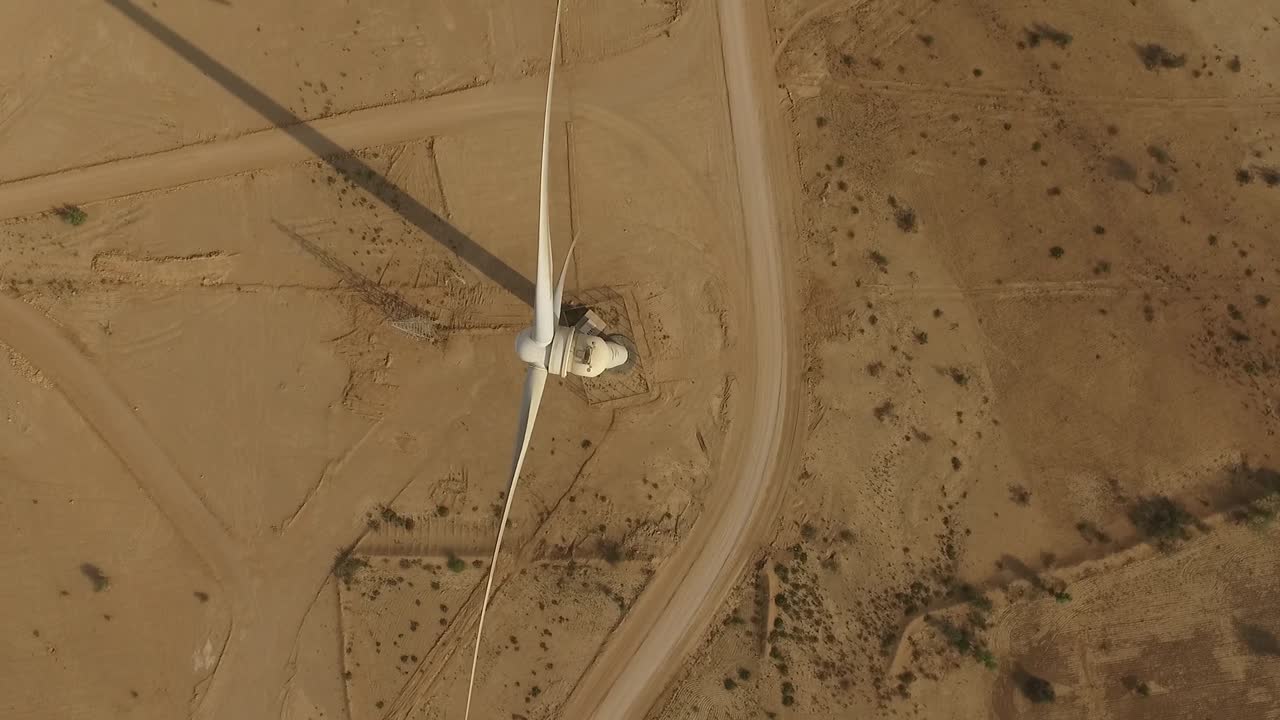 Drone view of a wind turbine in a dry, rugged desert. Long shadows, winding road, and sparse vegetation highlight renewable energy in remote, sunlit terrain