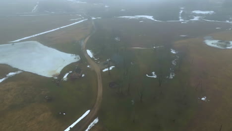 Aerial-footage-of-backcountry-farm-houses-amidst-scattered-trees-in-grassy-landscape,-foggy-climate-in-winter