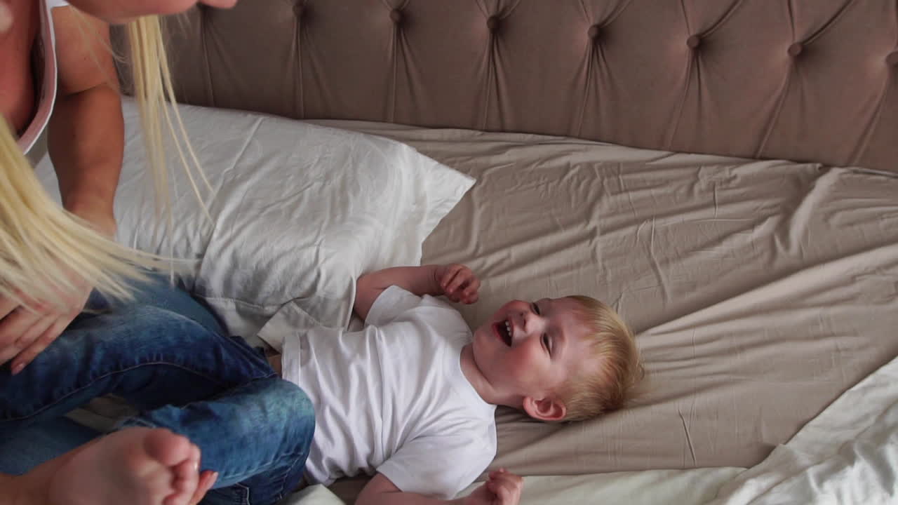 Mom tickles the boy lying on the bed who looks directly into the camera and laughs loudly