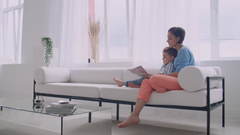 a young mother with a child reading a book sitting in a bright white interior of the house in the living room on the couch