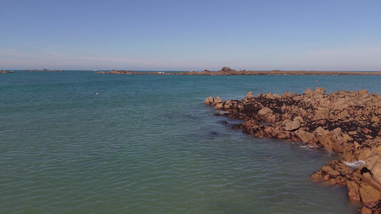 Isole del Canale di Guernsey. Filmato lento e molto basso con drone in avanti sopra formazioni rocciose con la bassa marea, sotto il sole del tardo pomeriggio, con mare calmo e cielo azzurro e viste su affioramenti rocciosi distanti all'orizzonte.