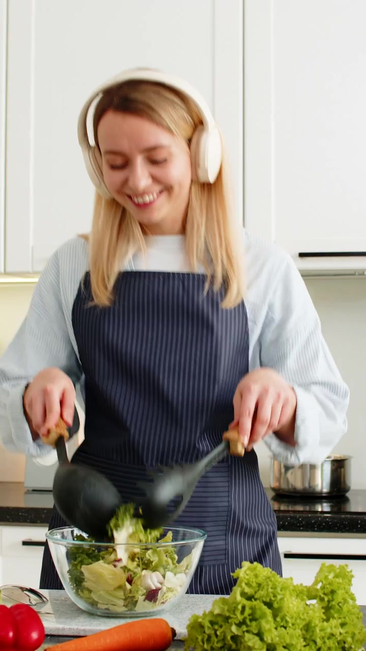 Młoda kobieta tańcząca w słuchawkach podczas mieszania świeżej sałatki warzywnej na dietę keto w domowej kuchni