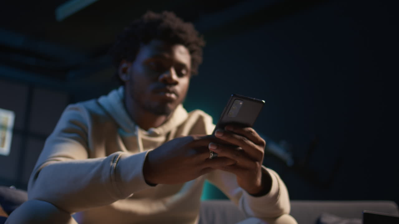 African American man scrolling through social media on smartphone while relaxing on a couch, cozy loft with neon lights. Person showcases comfort, online activity and digital engagement. Camera B.