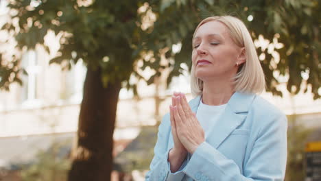 mature caucasian businesswoman standing on street city folding palms praying asking god for help