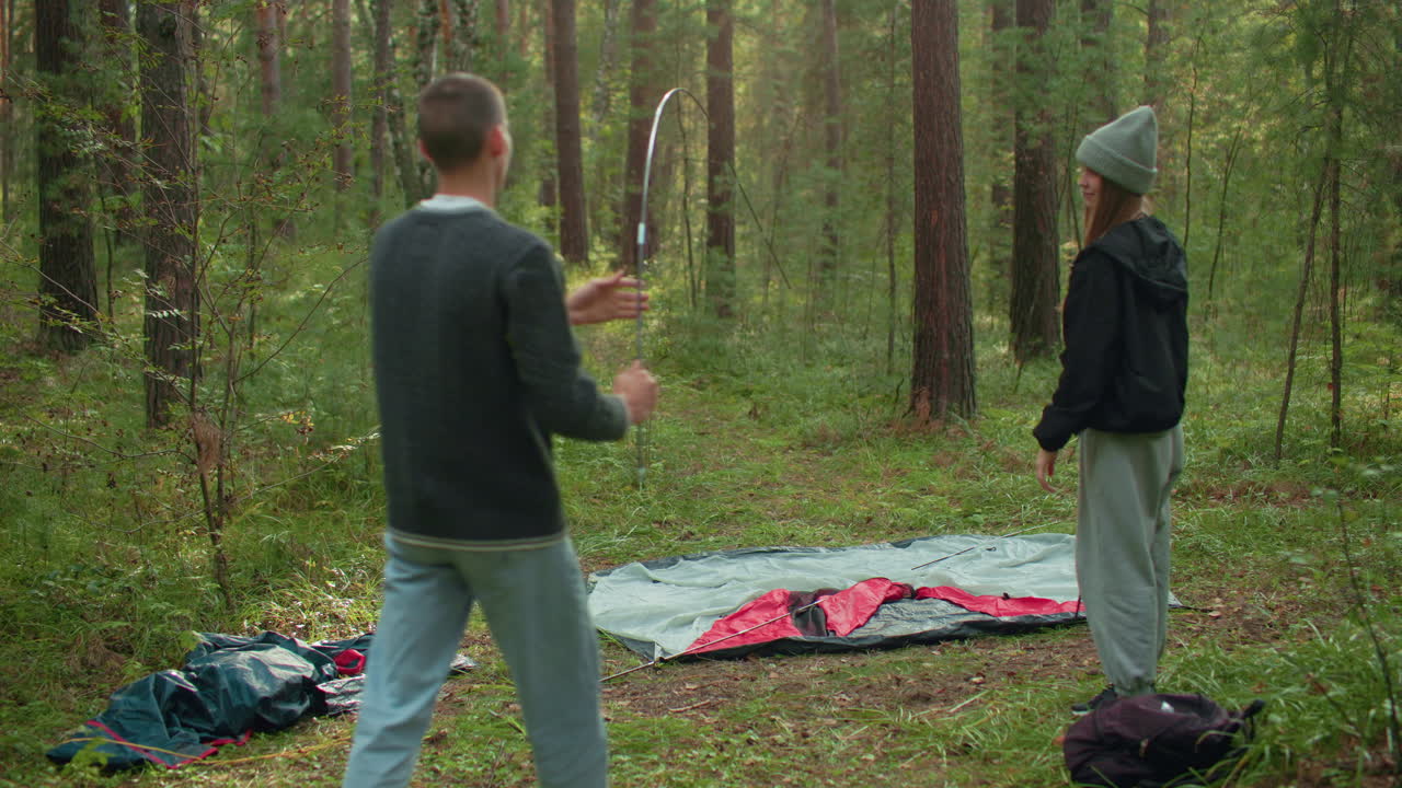 Young man stretches tent pole to ground aiming to fix it over tent fabric while lady stands nearby watching in quiet forest setting, surrounded by pine trees and scattered camping gear