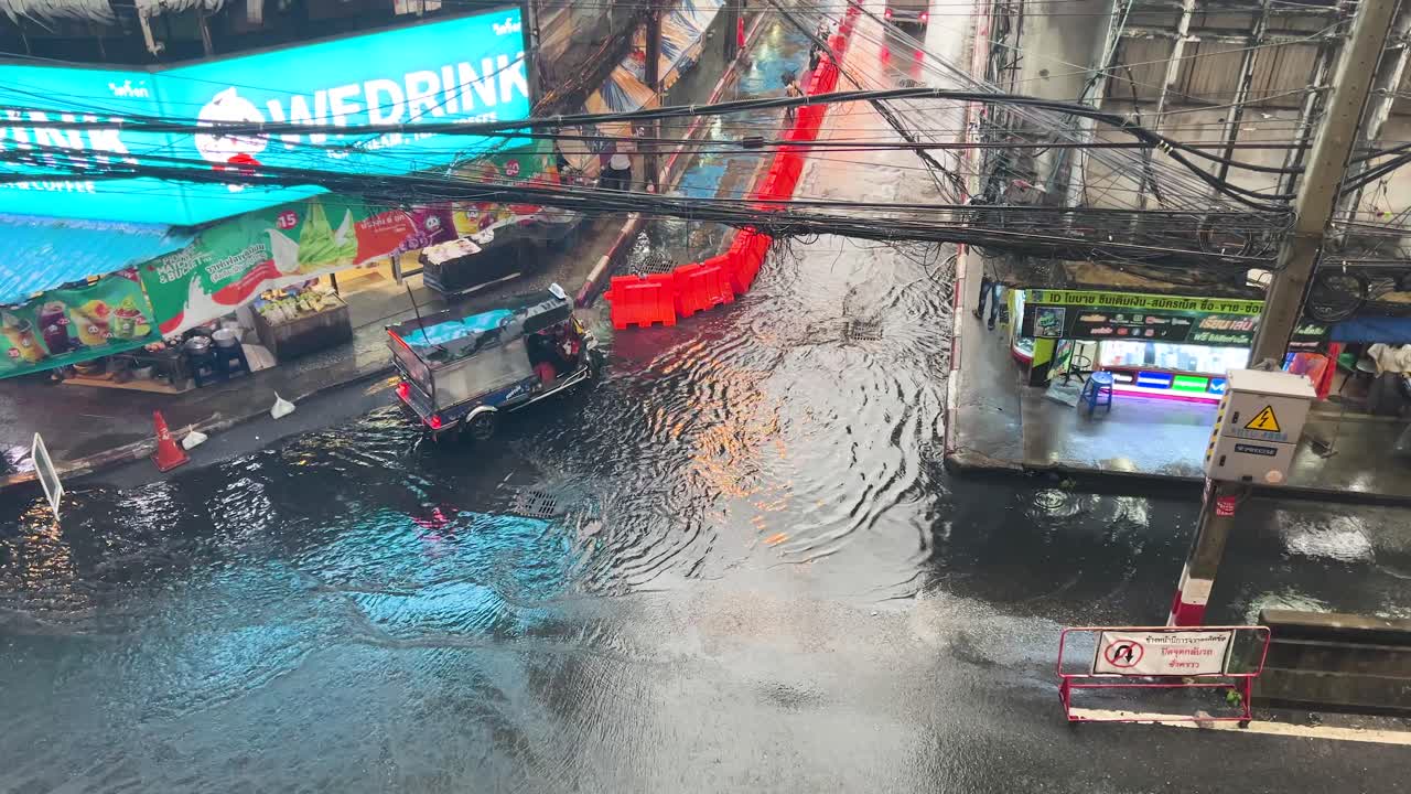 Tuk-tuks and motorbikes drive through a heavily flooded urban road in Bangkok, Thailand, under overcast daylight with visible reflections and slow-moving traffic