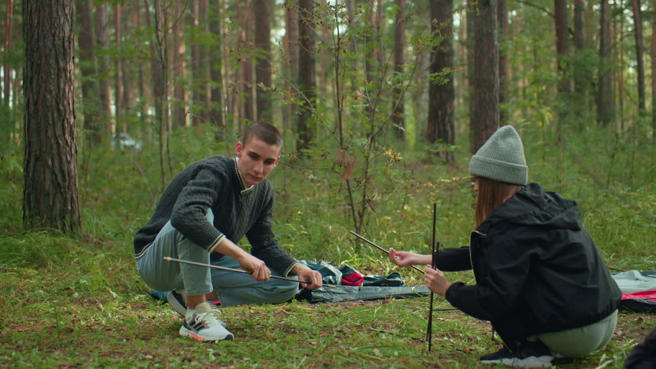 Brother and sister out camping in forest as sister holds tent pole waiting to pass next one to brother while he joins her to continue assembling tent surrounded by green grass and wooded background