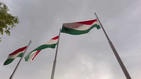 Tres-Banderas-Húngaras-En-Astas-De-Bandera-Que-Soplan-En-El-Viento-Con-Nubes-De-Tormenta-En-El-Fondo---Cámara-Lenta-De-120-Fps