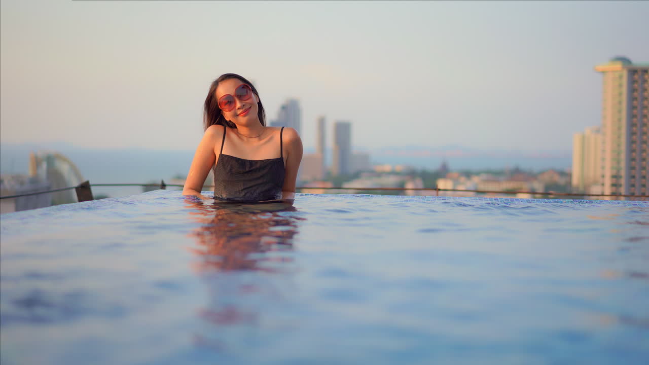 atractiva turista asiática feliz en la piscina infinita en la azotea de un exclusivo hotel resort, toma estática con espacio para copiar y horizonte urbano en segundo plano