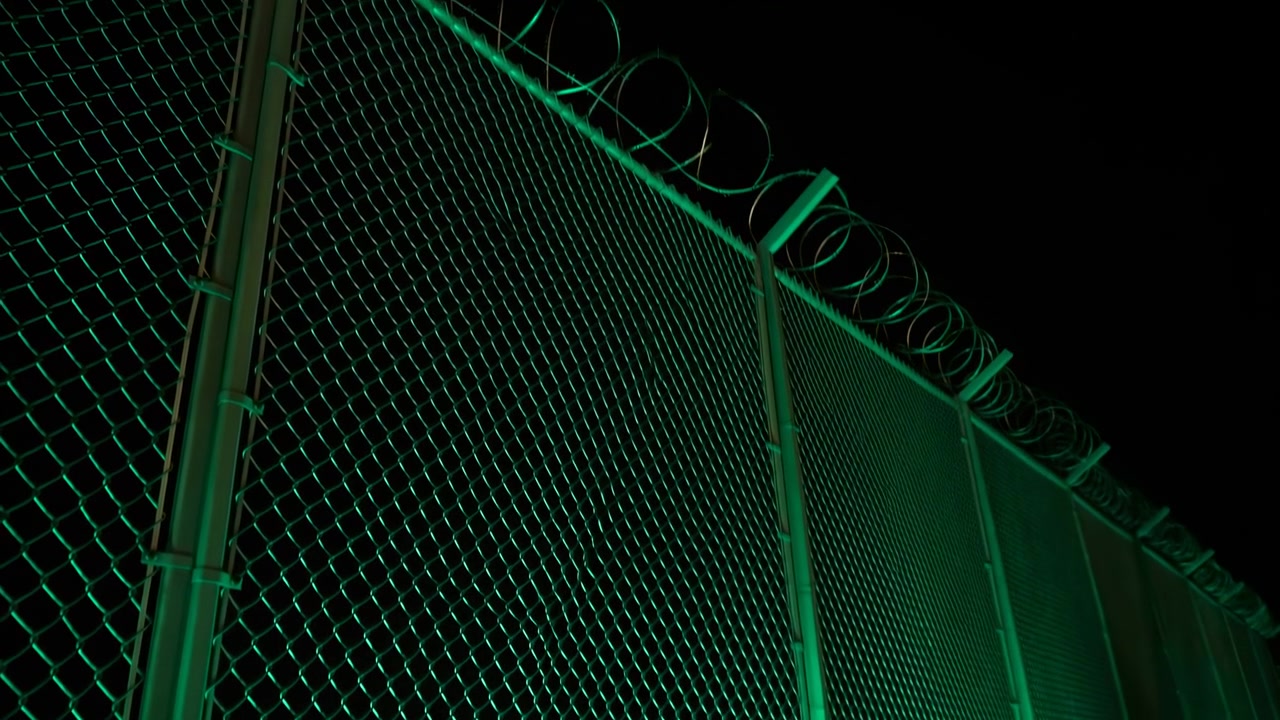 Una larga valla de seguridad metálica se extiende a lo lejos bajo una vívida iluminación verde, con alambre de púas en la parte superior y un fondo urbano oscuro