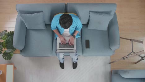 top view zoom out shot of a man typing on a laptop while sitting on the sofa with a smartphone at home