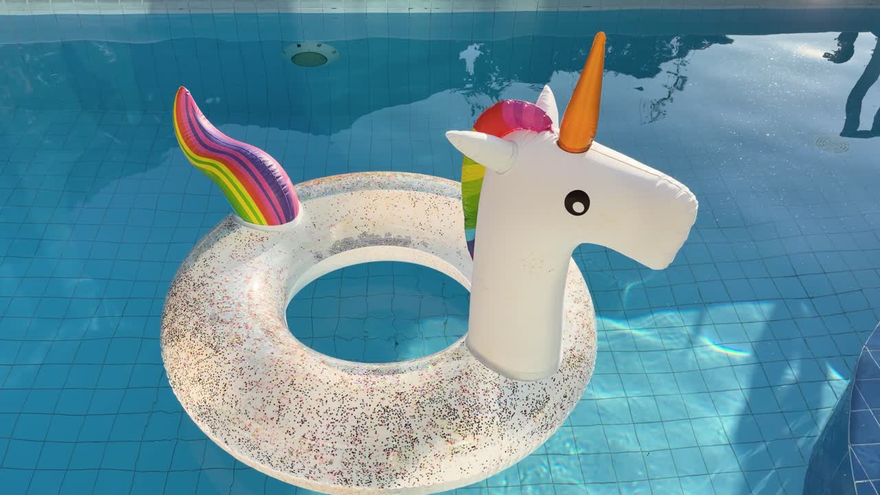 Colorful unicorn inflatable ring drifts on clear blue swimming pool, bright afternoon sunlight shimmering