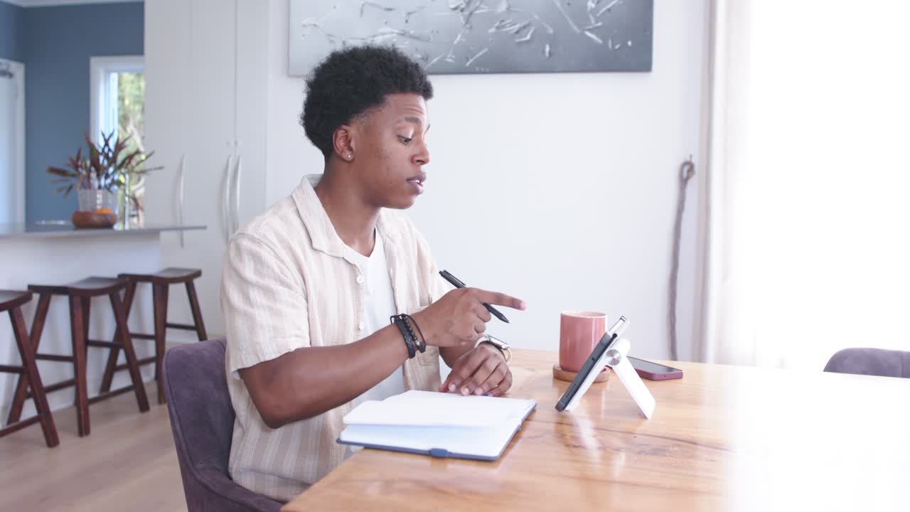 Writing man in short-sleeve shirt leaning at wooden table at home, pointing to tablet and notebook