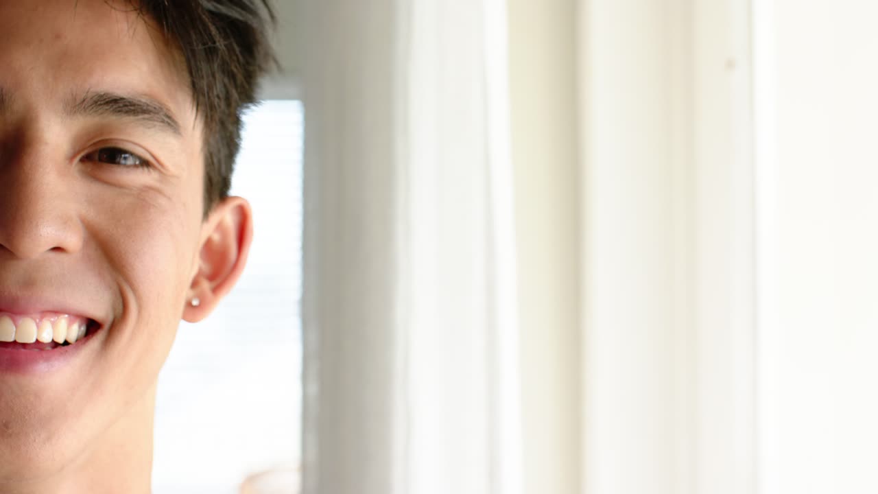 Opening clip showing Asian man smiling and holding gaze at home beside sheer curtains, copy space