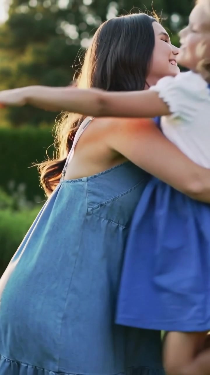 Video verticale: Adulto in abito blu che si protende per abbracciare un bambino con salopette blu che entra da destra, al parco