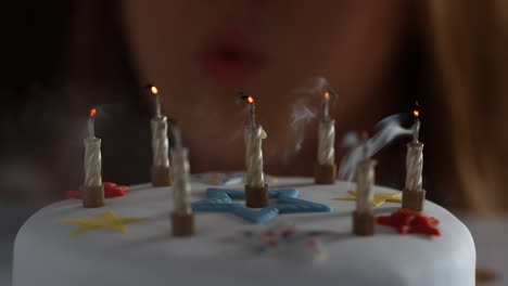 Close up of a woman blowing out seven lit candles on a white, decorated birthday cake, a party blower beside it, bokeh lights in the background, detail