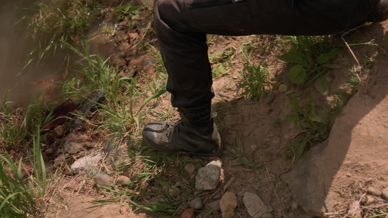 Partial view of soldier crouching while moving down grassy, uneven slope toward muddy riverbank, wearing tactical gloves and black boots, surrounded by dense vegetation under natural daylight outdoors