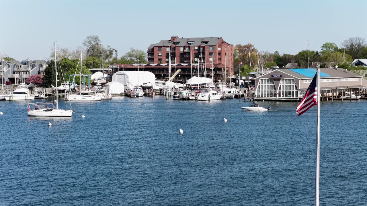 Vista aerea del porto di Annapolis, Maryland, con la bandiera americana che sventola sull'acqua calma, barche ed edifici del porto turistico sotto la luminosa luce solare primaverile, mostrando lo stile di vita costiero panoramico in una storica città degli Stati Uniti.