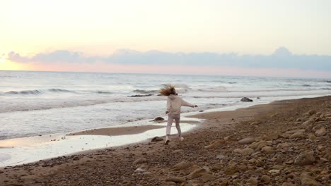 Happy-young-woman-with-long-blonde-hair-in-white-suit-running-along-ocean-beach-at-sunset