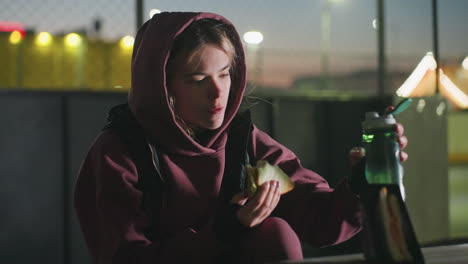 sportswoman in hoodie sits outdoors at dusk under bright city lights eating sandwich while sipping water from bottle beside snack tray on wooden table in urban park breathing crisp cool air