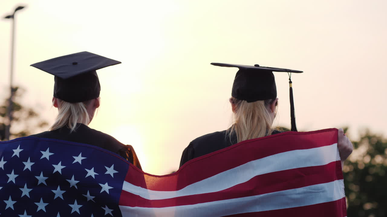 dos mujeres graduadas universitarias levantan las manos sobre los hombros de la bandera de estados unidos