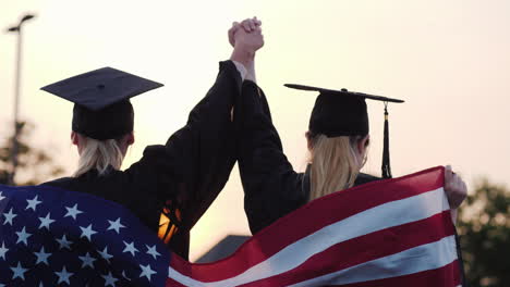 two women college graduates raise their hands up on the shoulders of the usa flag