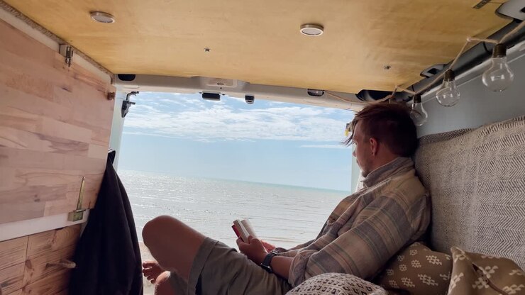 Giovane uomo caucasico in un furgone di fronte al mare che legge pensieroso.