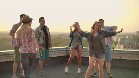 A-company-of-six-young-people-threw-a-party-on-the-roof-with-beer.-Three-young-men-and-two-young-girls-are-dancing-in-plaid-shirts.-Young-blonde-dances-in-the-foreground-in-denim-shorts-and-denim-jacket.