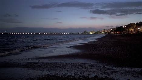 ground level footage of seashore at kalamata beach during dusk, calming waves on sea, city and pier lights on the background, static shot 4k