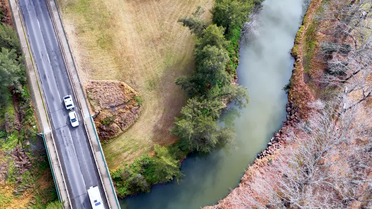 Drone footage tracks vehicles driving across a two-lane bridge spanning a winding river in a rural, grassy landscape with scattered trees and soft daylight